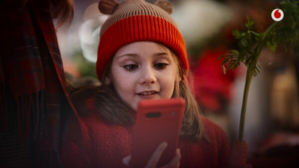 Vodafone is celebrating the power of connection in its new Christmas advert featuring a hero elf and a young girl's scheme to capture Santa on camera, depicting a young girl in a red and brown had looing at a phone with a red case