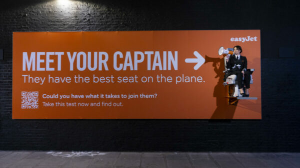 Text on a bright orange billboard, the same bold colour as easyJet's branding, reads "Meet your Captain. They have the best on the plane". It then points to Captain Sarah Ackerley who is dressed in her uniform holding a megaphone and is sat on an elevated pilot seat, which is part of the billboard. Underneath text next to a QR code reads "Could you have what it takes to join them? Take this test now and find out."