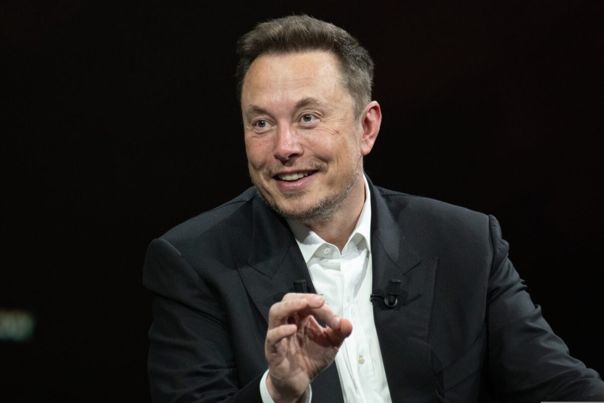 Elon Musk depicted smiling whilst explaining something to an audience, dressed smartly in a black suit. X slammed as it reintroduces blue ticks