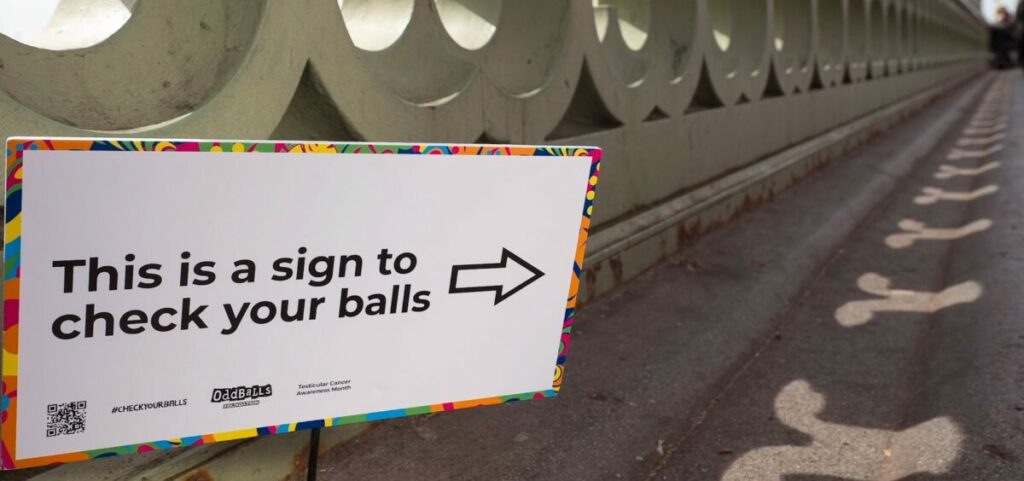 Image shows Westminster Bridge with phallic shadows appearing as the sun shines through the bridge. A sign reads "This is a sign to check your balls". Charity the Oddballs foundation has turned London's Westminster Bridge into an intelligent installation for Testicular Cancer Awareness Month.