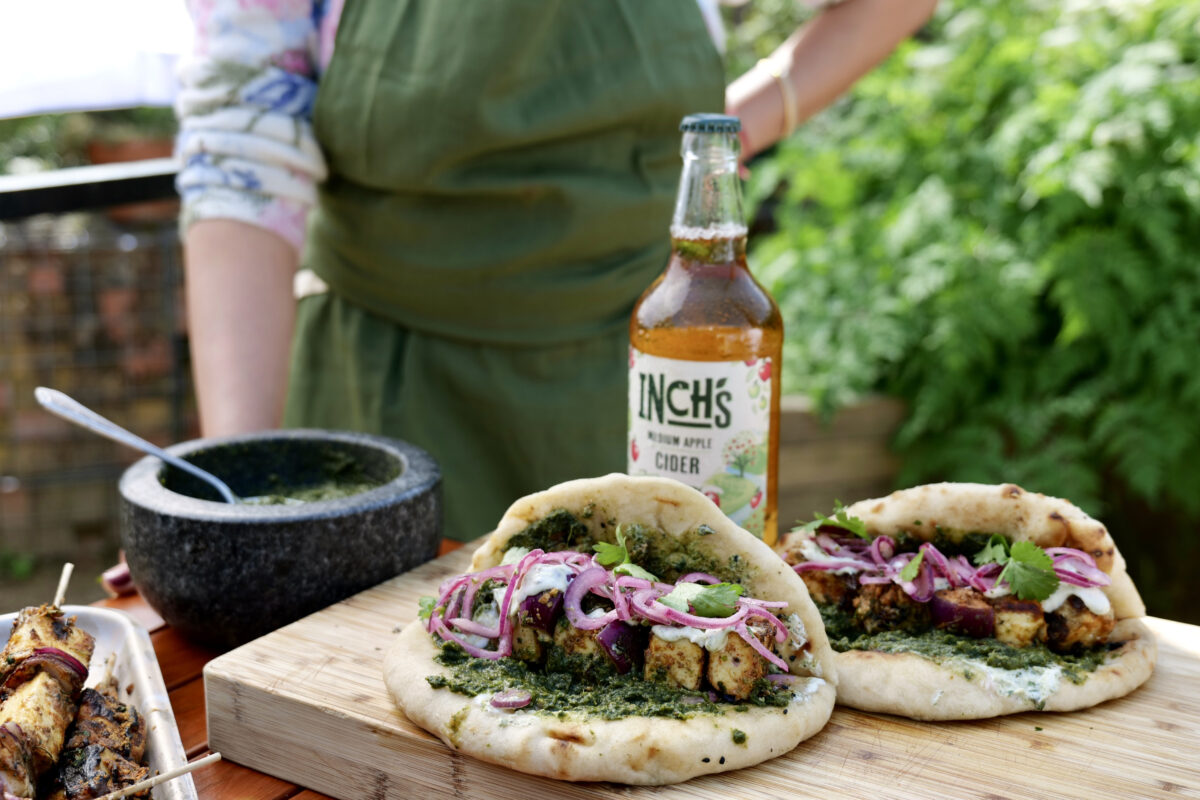 Chef stands in a green apron in front of an outside table in a pretty garden with a chopping boar and wraps on flat bread and a bottle of Inch's. Heineken UK brand Inch's Cider is teaming up with leading food influencers including Big Has, Alfie Cooks and Hey Renu to show off a set of locally sourced recipes featuring the cider.