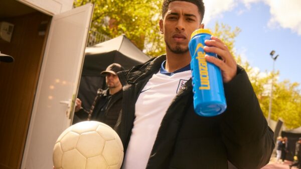 Jude Bellingham poses holding a Lucozade bottle and a football while wearing an England shirt. Lucozade has brought on board Jude Bellingham in its newest sponsorship deal, with Bellingham to feature in a high energy social post.