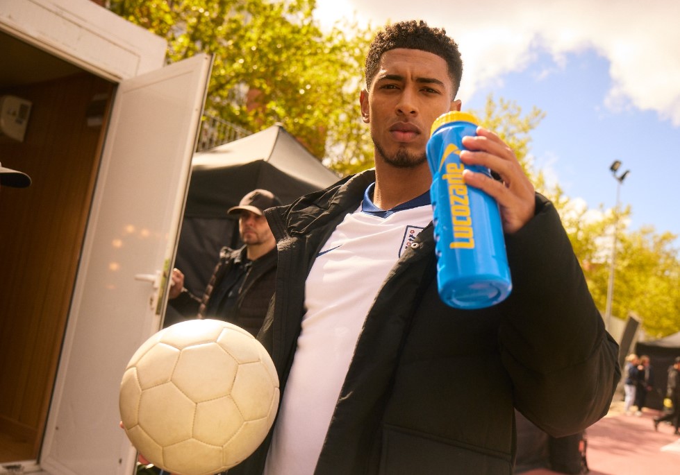 Jude Bellingham poses holding a Lucozade bottle and a football while wearing an England shirt. Lucozade has brought on board Jude Bellingham in its newest sponsorship deal, with Bellingham to feature in a high energy social post.