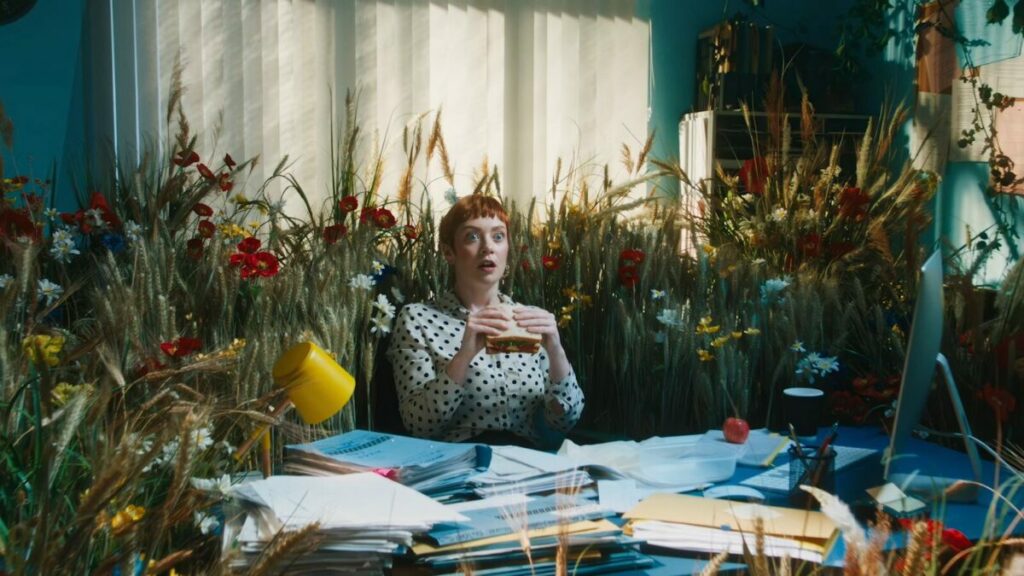 Woman munches on sandwich made with Wildfarmed bread at her desk, she is suddenly transported into a floral fantasy with pink, long verdant flowers. Regenerative food and farming company Wildfarmed is celebrating the launch of its 'life-changing' bread range in its first major advertising campaign.