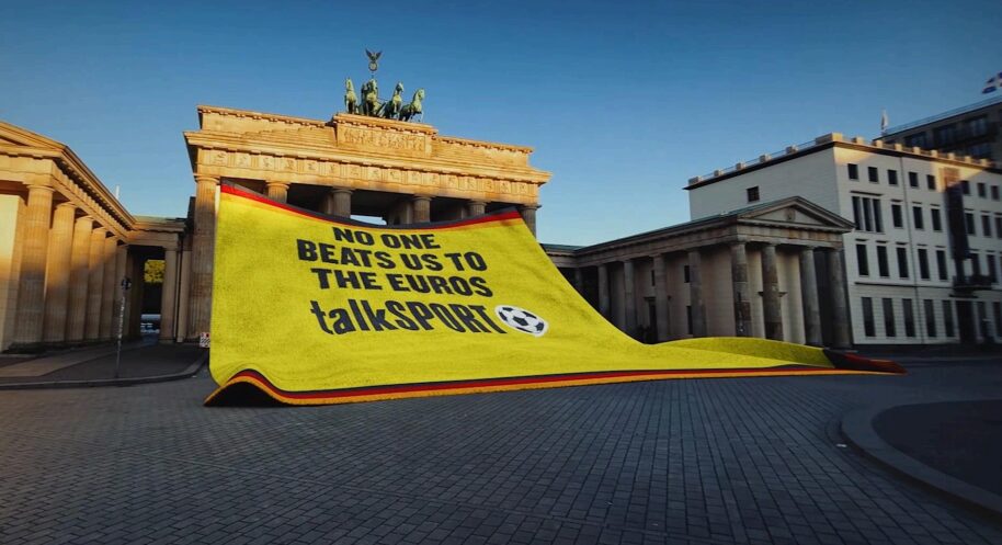 A towl yellow tih a black red yellow border like the German flag drapes over the Brandenburg gate on a sunny Berlin day. It read "No one beats us to the Euros" and it features talksport's little football logo. Talksport has launched its Euro 2024 campaign with a cheeky dig at the Germans, in a spot which opens with a shot of the Brandenburg gate and a towel that reads "No one beats us to the Euros".