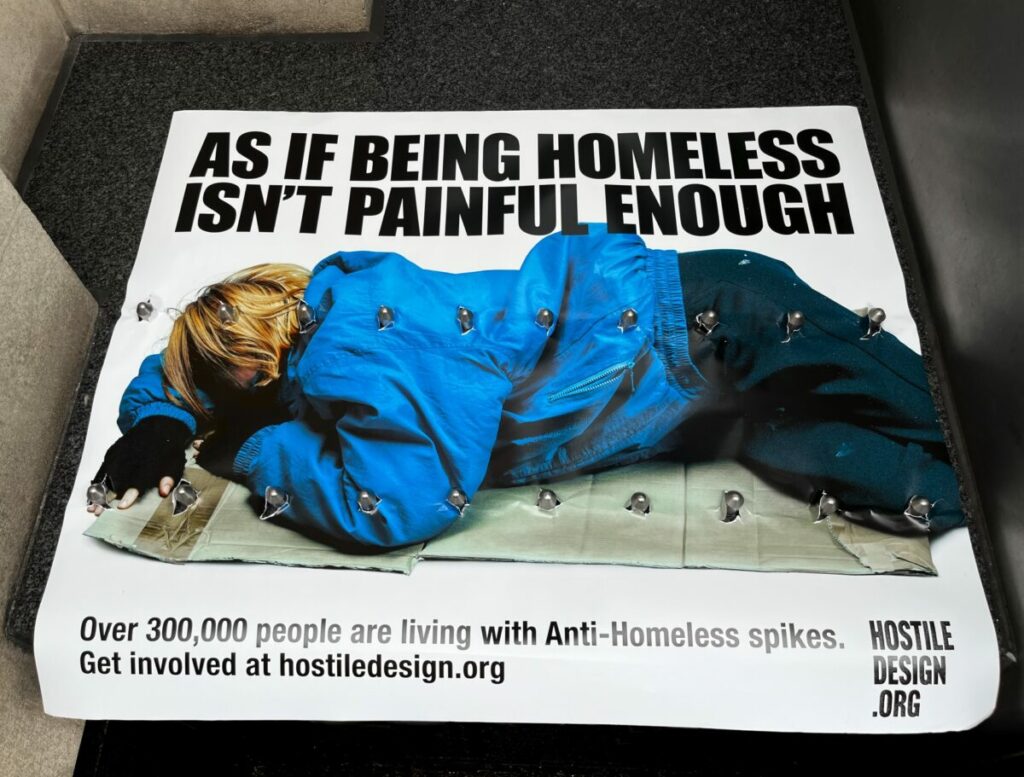 Spikes poke through a banner beside a shop window. The banner shows a the words "Welcome to the sharp end of homelessness". It highlights the piercing impact of hostile architecture on those who sleep rough.
