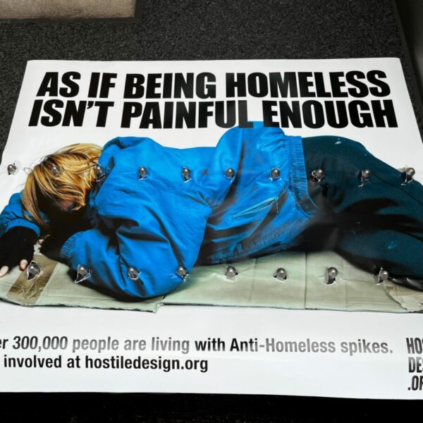 Spikes poke through a banner beside a shop window. The banner shows a the words "Welcome to the sharp end of homelessness". It highlights the piercing impact of hostile architecture on those who sleep rough.