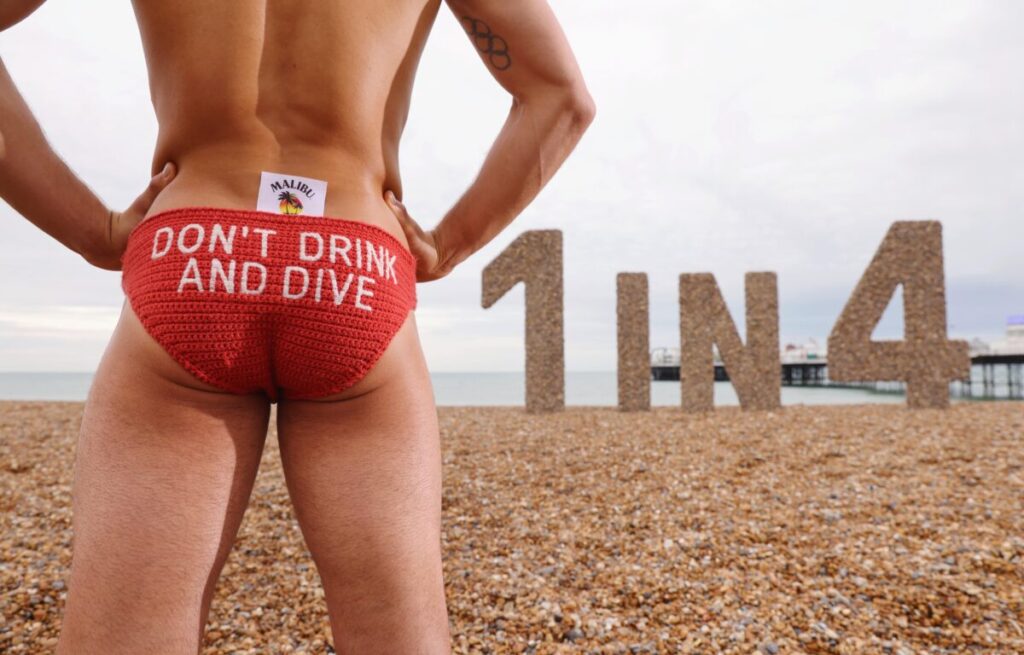 Tom Daley wears a pear of knitted speedo shaped trunks with the text "Don't drink and dive' as part of a tie-up with the Royal Life Saving Society. From the brand's latest Tom Daley spot, to its collaborations with Oatly and Aries Malibu's Craig van Niekerk shares ingredients to the perfect collab. Stands in front of a giant 1 in 4 statue made from stones on Brighton beach.