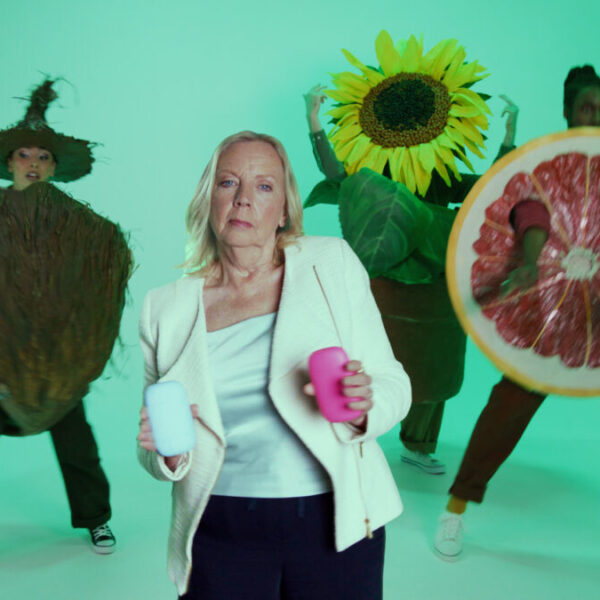 Deborah Meaden holds up her Fussy deodorant against a green backdrop with fun alien like figures. Natural deodorant brand Fussy shares a 90s-style rap battle video complete with a stern looking Deborah Meaden showing off her dance moves.